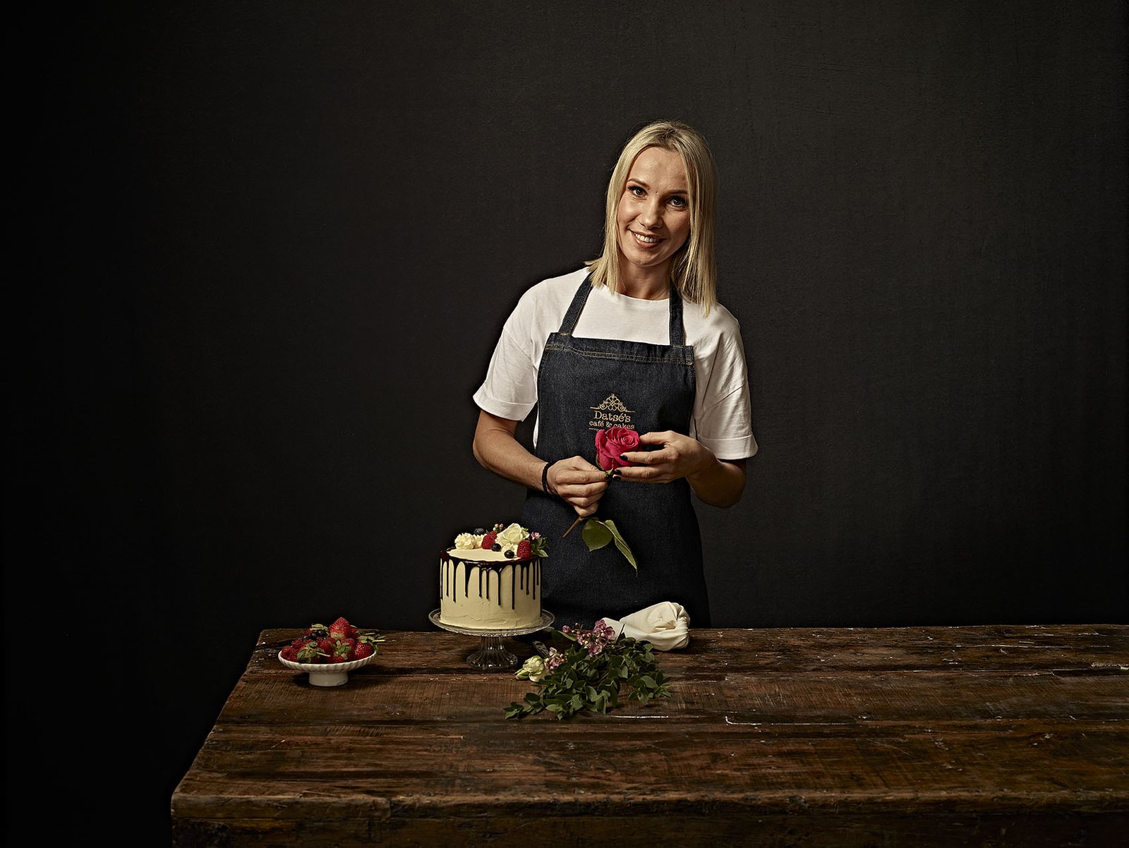 The baker at Datsé’s Café and Cakes, in the shop apron, with a berry drip cake and a rose